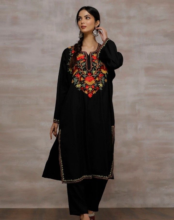 Woman wearing a black Kashmiri phiran (traditional long tunic) with vibrant floral embroidery in red, orange, and yellow on the chest area. The outfit features intricate gold trim details along the neckline and hem. She's paired it with matching black pants and traditional jewelry including ornate earrings. The model is posed elegantly against a neutral beige textured background.