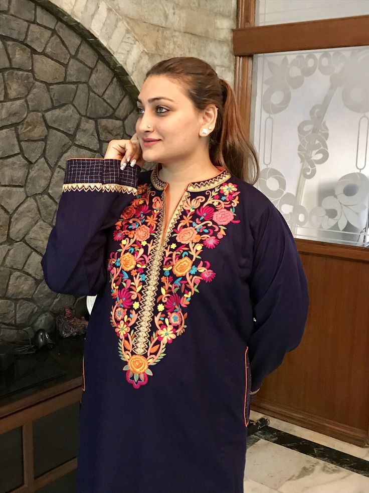 Woman in elegant navy blue traditional kurta featuring intricate multicolored floral embroidery with pink, orange, and gold thread work around the neckline and front panel, posed in an upscale interior setting