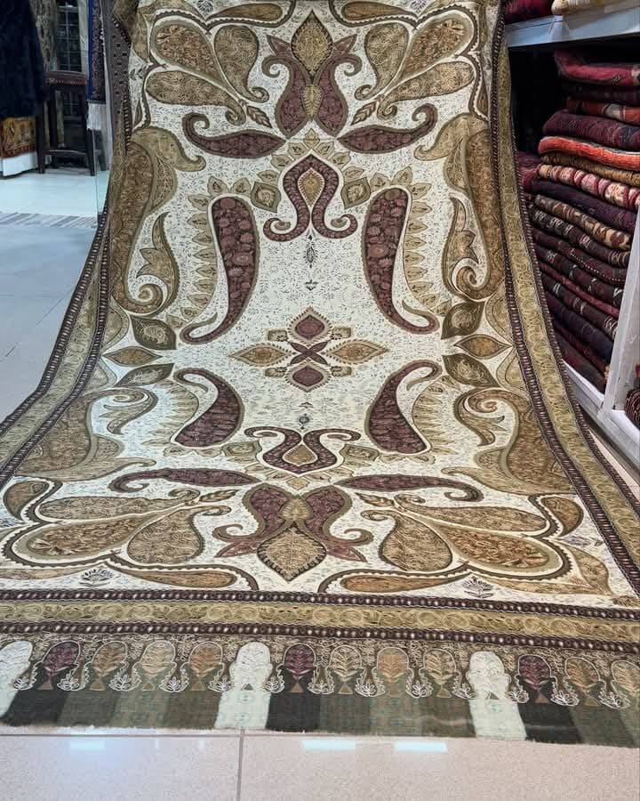 Luxurious handwoven Pashmina shawl with intricate paisley and floral patterns in rich earth tones of cream, gold, and burgundy, displayed in a textile shop with traditional ornate border design and decorative fringe